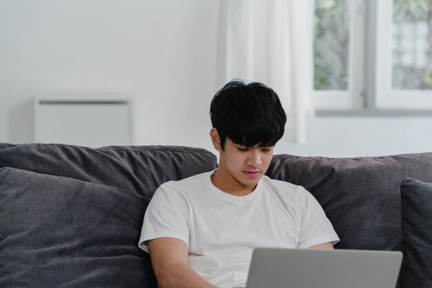 Hombre asiático independiente que trabaja en casa, hombre creativo en la computadora portátil en el sofá en la sala de estar. Empresario joven empresario empresario, jugar a la computadora, comprobar las redes sociales en el lugar de trabajo en la casa moderna.