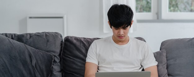 Hombre asiático independiente que trabaja en casa, hombre creativo en la computadora portátil en el sofá en la sala de estar. Empresario joven empresario empresario, jugar a la computadora, comprobar las redes sociales en el lugar de trabajo en la casa moderna.