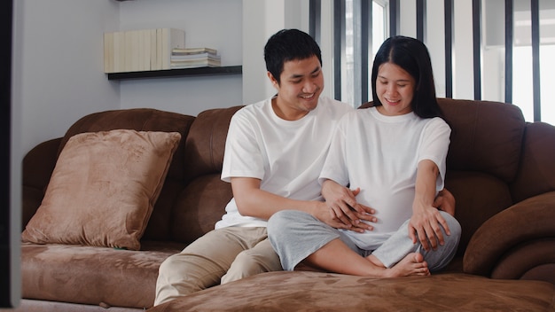 El hombre asiático joven de la pareja embarazada toca el vientre de su esposa hablando con su hijo. Mamá y papá se sienten felices sonriendo pacíficos mientras cuidan bebé, embarazo acostado en el sofá en la sala de estar en casa.