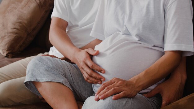 El hombre asiático joven de la pareja embarazada toca el vientre de su esposa hablando con su hijo. Mamá y papá se sienten felices sonriendo pacíficos mientras cuidan bebé, embarazo acostado en el sofá en la sala de estar en casa.