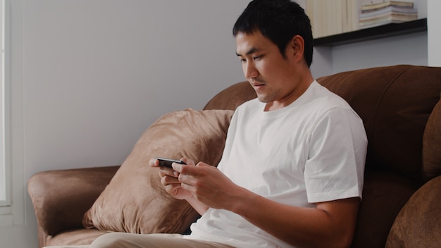 Hombre asiático joven que usa el teléfono móvil que juega a videojuegos en la televisión en la sala de estar, varón que se siente feliz usando el tiempo de la relajación que miente en el sofá en casa. Los hombres juegan juegos de relax en casa.