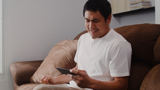 Hombre asiático joven que usa el teléfono móvil que juega a videojuegos en la televisión en la sala de estar, varón que se siente feliz usando el tiempo de la relajación que miente en el sofá en casa. Los hombres juegan juegos de relax en casa.