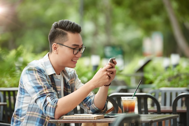 Hombre asiático joven vestido informalmente sentado en un café al aire libre y con smartphone