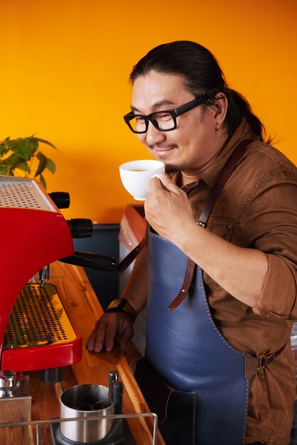 Hombre asiático de mediana edad en delantal de pie junto a la máquina de café y sosteniendo la taza a la nariz