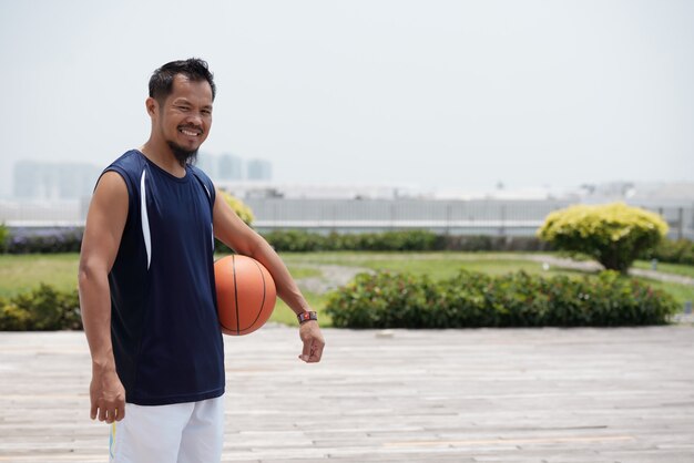 Hombre asiático de pie al aire libre en el estadio, sosteniendo baloncesto y sonriendo