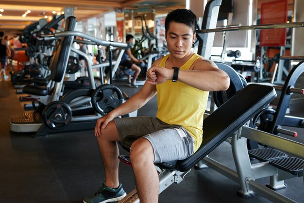 Hombre asiático sentado en el banco en el gimnasio y mirando el reloj de pulsera