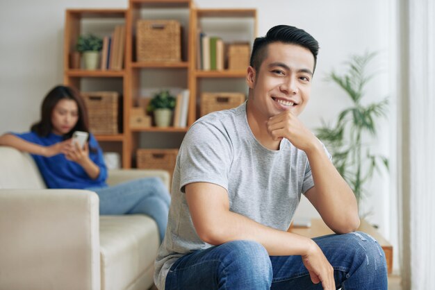 Hombre asiático sentado en su casa con la barbilla en la mano, y una mujer con un teléfono inteligente detrás de él en el sofá