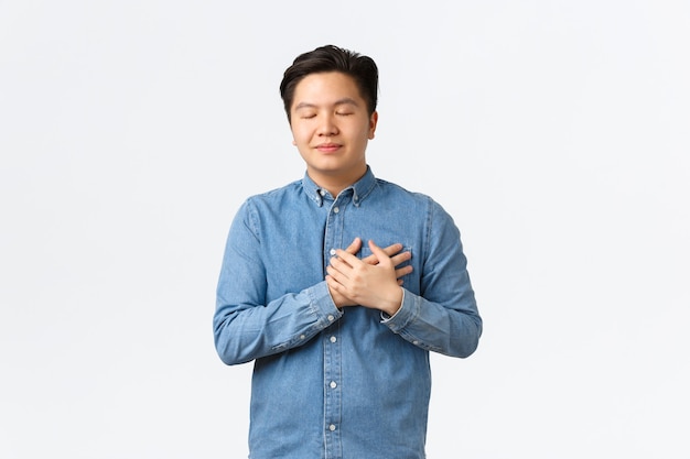 Hombre asiático soñador esperanzado en camisa, ojos cerrados y tomados de la mano en el corazón, imaginando algo, sintiendo amor y cuidado, recordando un buen recuerdo, pensando en alguien, fondo blanco