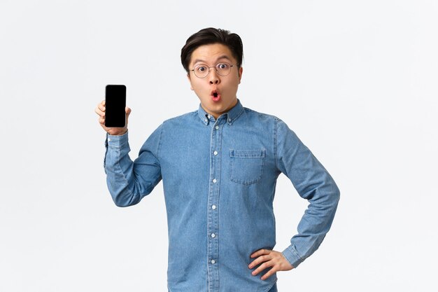 Hombre asiático sorprendido y asombrado con gafas y ropa casual, mostrando la pantalla del teléfono móvil y diciendo wow, discutiendo la nueva aplicación de teléfono inteligente, últimas noticias en línea, fondo blanco