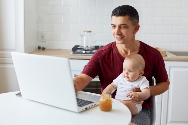 Hombre de aspecto agradable con camiseta casual marrón, padre adulto joven sentado a la mesa en la cocina frente a la computadora portátil mirando la pantalla del portátil con expresión positiva.