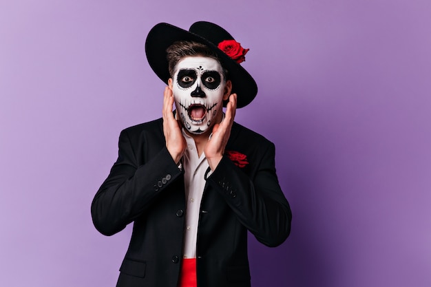 Hombre asustado con sombrero de ala ancha mirando con horror a la cámara. Retrato de chico con maquillaje de Halloween posando sobre fondo morado.