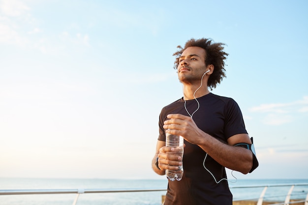 Hombre atleta bebiendo agua de una botella de plástico después de un duro entrenamiento. Deportista masculino de piel oscura mirando al cielo mientras corre, disfrutando de la vista