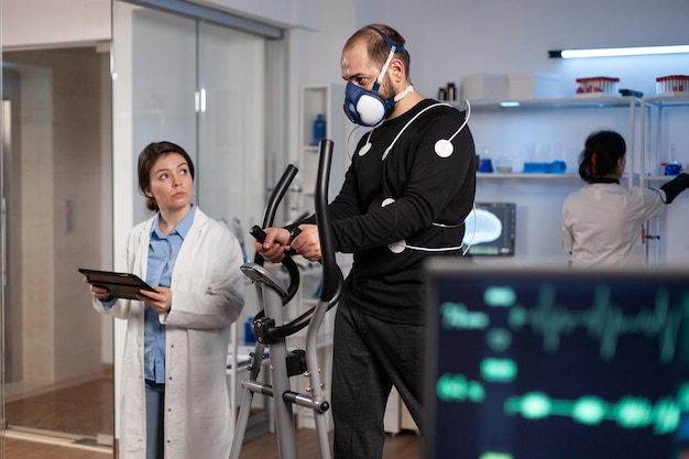 Hombre atleta con electrodos médicos corriendo en bicicleta de fitness trabajando en la resistencia corporal durante el entrenamiento médico en un laboratorio moderno mientras el médico monitorea los datos de EGK. Pulso de frecuencia cardíaca en el monitor