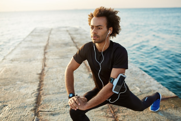 Hombre atleta en ropa deportiva negra estirando las piernas con estocada ejercicio de estiramiento de isquiotibiales en el muelle. Escuchar música en auriculares.