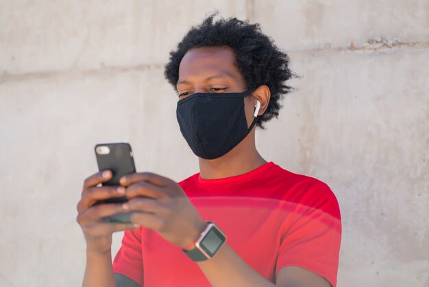 Hombre atlético afro con mascarilla y usando su teléfono móvil después de hacer ejercicio al aire libre. Concepto de deportes y tecnología. Nuevo estilo de vida normal.