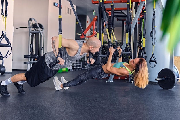 Hombre atlético de cabeza rapada y mujer morena delgada haciendo ejercicio con correas trx en un club de gimnasia.