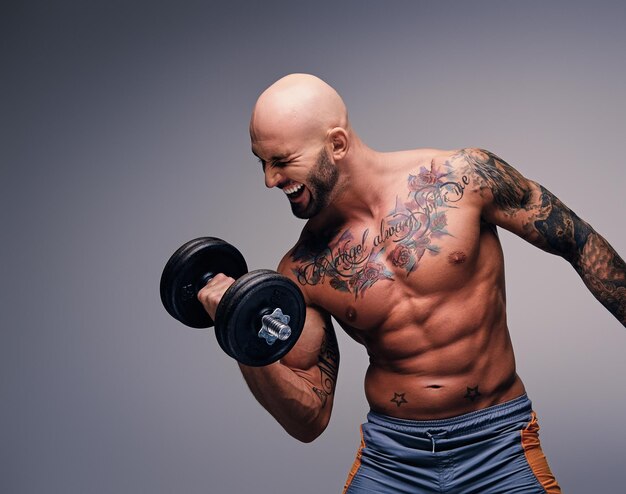 Hombre atlético de cabeza rapada con tatuajes en el torso y los brazos sostiene pesas aisladas en el fondo de la viñeta gris.