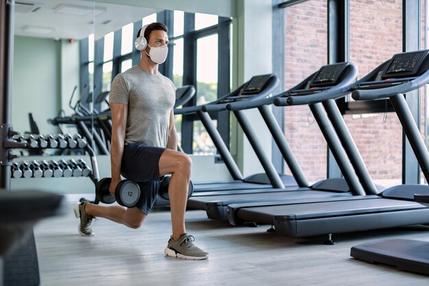 Hombre atlético con mascarilla que entrena con pesas en posición de estocada en un gimnasio