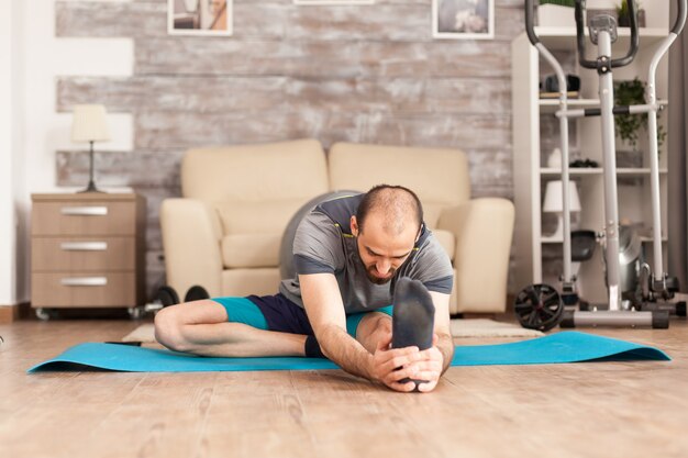 Hombre atlético que se extiende antes del entrenamiento en la estera de yoga en casa durante la pandemia global.