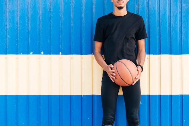 Foto gratuita hombre atlético sin rostro de pie contra la pared sosteniendo baloncesto