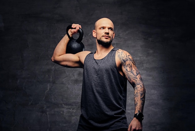 Hombre atlético tatuado con la cabeza rapada haciendo ejercicios de hombros con pesas rusas.