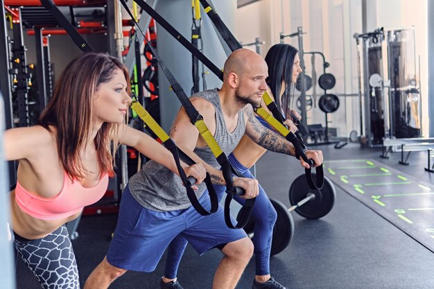 Hombre atlético tatuado y dos mujeres deportivas haciendo ejercicios de correas trx.