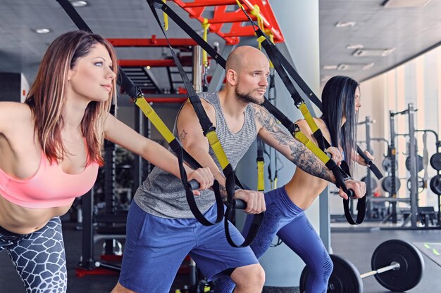 Hombre atlético tatuado y dos mujeres deportivas haciendo ejercicios de correas trx.