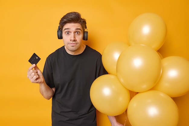 Un hombre atónito se prepara para la celebración vestido con una camiseta negra informal que sostiene una tarjeta bancaria y un montón de globos escucha la pista de audio a través de las poses de los auriculares contra el fondo amarillo Hora de la fiesta