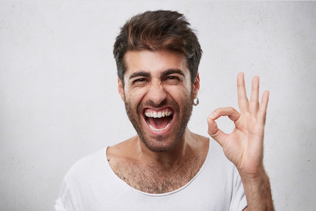 Hombre atractivo con barba cerrando los ojos y abriendo la boca con alegría mostrando el signo de ok estar contento