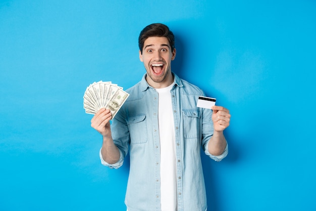 Hombre atractivo feliz mirando sorprendido, mostrando efectivo y tarjeta de crédito, concepto de bancos, crédito y finanzas. Fondo de estudio azul.