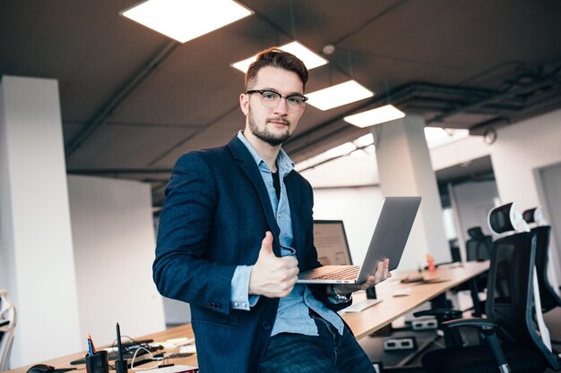 Hombre atractivo en glassess está de pie cerca del lugar de trabajo en la oficina. Viste camisa azul, chaqueta oscura. Sostiene la computadora portátil, muestra bien la señal y mira a la cámara.