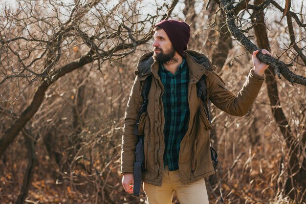 Hombre atractivo hipster viajando con mochila en el bosque de otoño con chaqueta y sombrero, turista activo, explorando la naturaleza en la estación fría