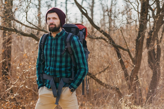 Hombre atractivo joven inconformista viajando con mochila en el bosque de otoño con camisa a cuadros y sombrero, turista activo, explorando la naturaleza en la estación fría