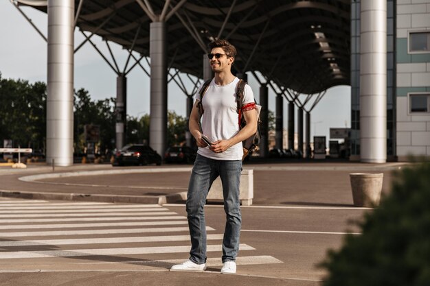 Hombre atractivo joven en jeans camiseta blanca y gafas de sol tiene pasaporte y mochila Chico morena de buen humor se mueve en la ciudad