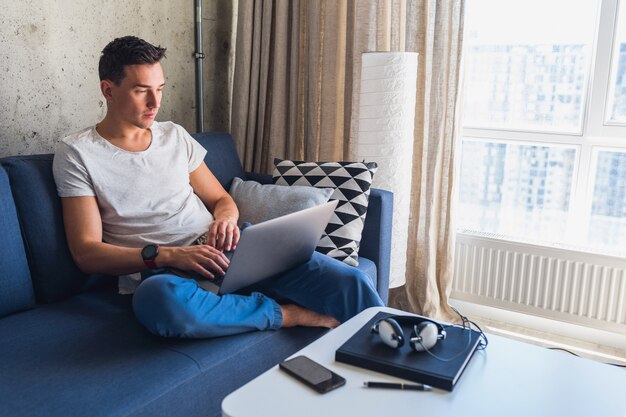 Hombre atractivo joven sentado en el sofá en casa trabajando con un portátil en línea, usando internet