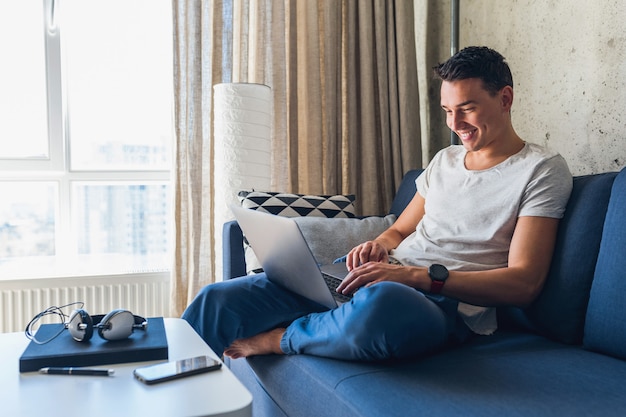 Hombre atractivo joven sentado en el sofá en casa trabajando con un portátil en línea, usando internet