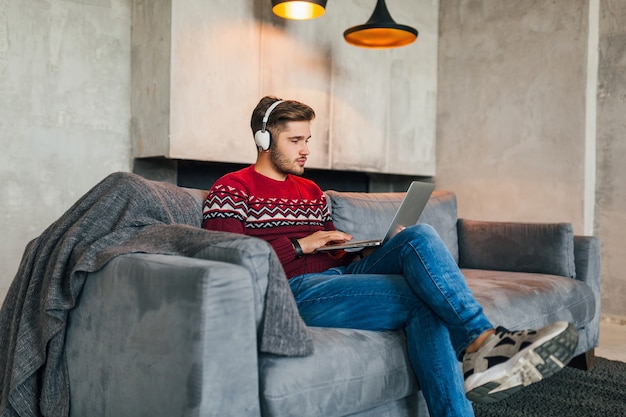 Hombre atractivo joven en el sofá en casa en invierno en auriculares, escuchando música, vistiendo un suéter de punto rojo, trabajando en una computadora portátil, autónomo, serio, ocupado, escribiendo, concentrado