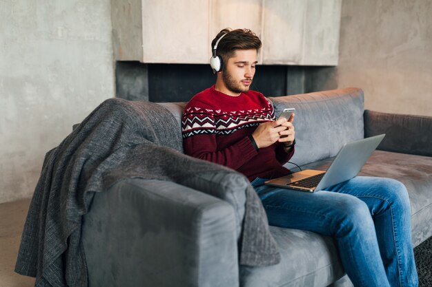 Hombre atractivo joven en el sofá en casa en invierno con el teléfono inteligente en los auriculares, escuchando música, vistiendo suéter de punto rojo, trabajando en la computadora portátil, autónomo