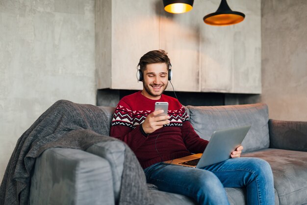 Hombre atractivo joven en el sofá en casa en invierno con el teléfono inteligente en los auriculares, escuchando música, vistiendo suéter de punto rojo, trabajando en la computadora portátil, autónomo