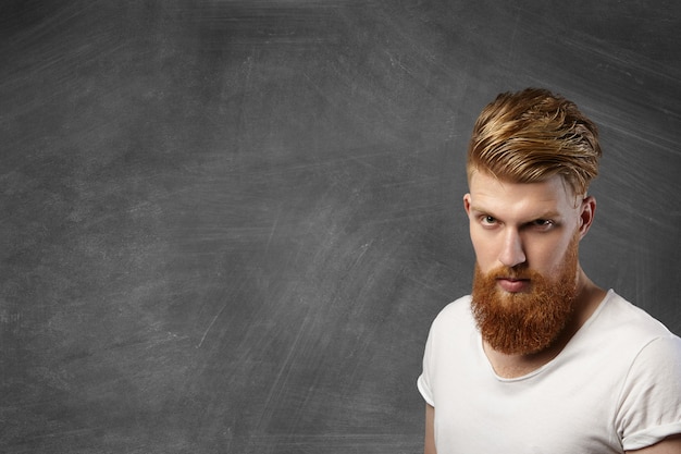 Foto gratuita hombre atractivo pelirrojo hipster con corte de pelo elegante y barba gruesa posando contra la pizarra en blanco con espacio de copia de su texto o contenido promocional, mirando con enojo.