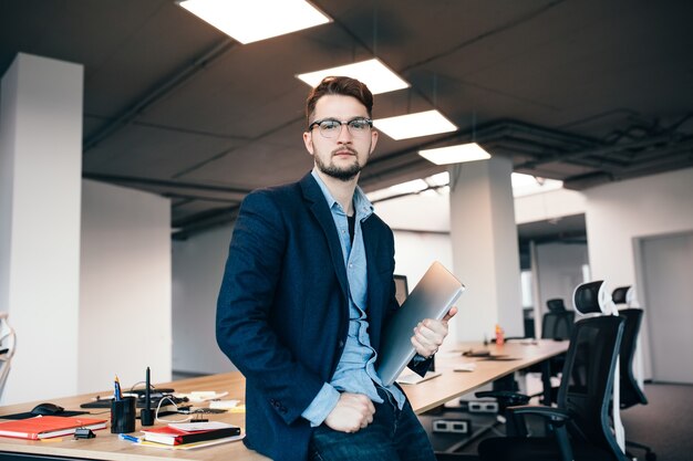 Hombre atractivo serio en glassess está de pie cerca del lugar de trabajo en la oficina. Viste camisa azul, chaqueta oscura, computadora portátil en la mano. Él está mirando a la cámara.