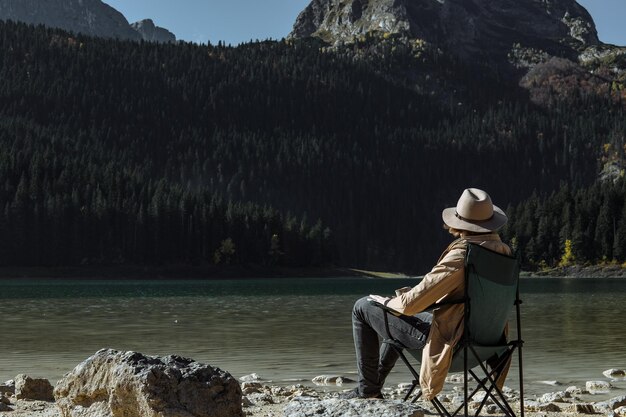 El hombre atractivo en el sombrero está bebiendo café y disfrutando de la vista del Parque Nacional Autumn Black Lake Durmitor Zabljak Montenegro