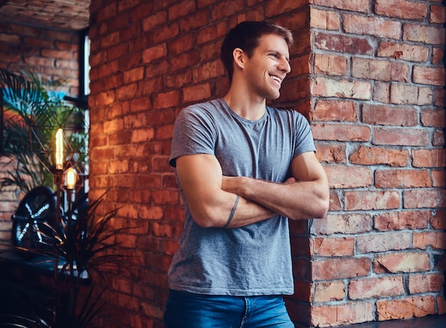 Foto gratuita un hombre atractivo tatuado con estilo vestido con jeans y una camiseta, se inclina contra la pared de ladrillo, con los brazos cruzados. sonriendo y mirando a otro lado.