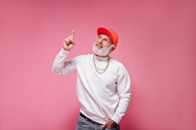 Hombre atractivo en traje elegante apunta hacia arriba sobre fondo rosa Chico elegante con cabello blanco en gorra naranja con cadena de oro posando