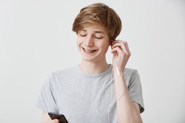 Hombre con auriculares blancos, sonriendo ampliamente, usando la aplicación de música, sosteniendo un teléfono inteligente moderno con conexión a Internet de alta velocidad, enviando mensajes de texto a sus amigos. Tecnología moderna y comunicación.