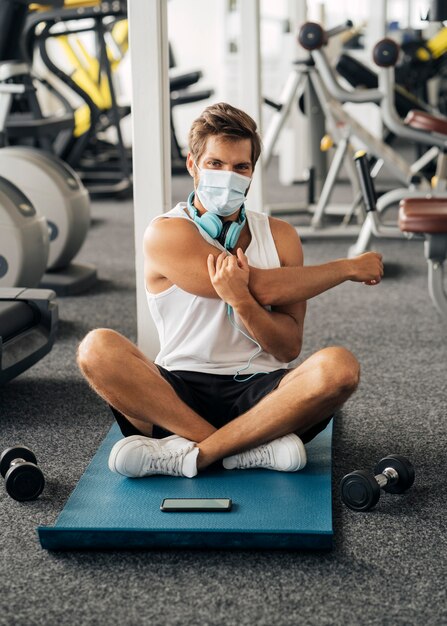 Hombre con auriculares y máscara médica en el gimnasio trabajando en mat