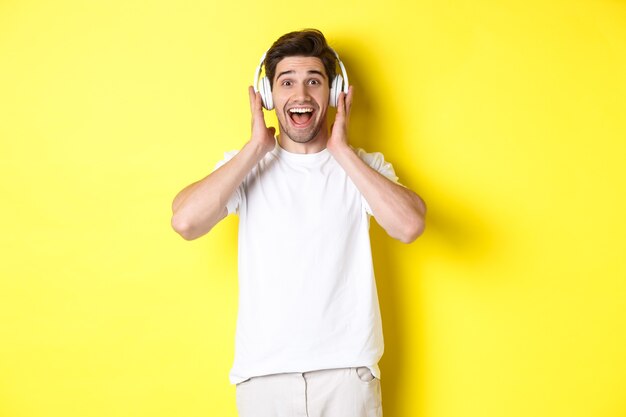 Hombre en auriculares mirando sorprendido y feliz, escuchando una canción impresionante, de pie sobre fondo amarillo.
