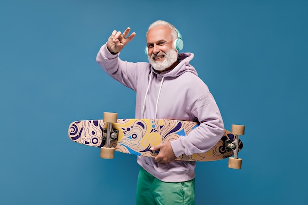 Hombre en auriculares mostrando el signo de la paz y sosteniendo una tabla larga Chico alegre con cabello gris en ropa elegante sonriendo sobre fondo aislado