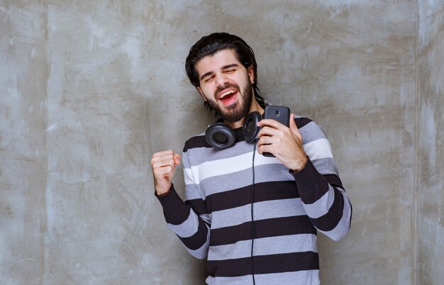 Hombre con auriculares sosteniendo un teléfono negro y mostrando signos de satisfacción