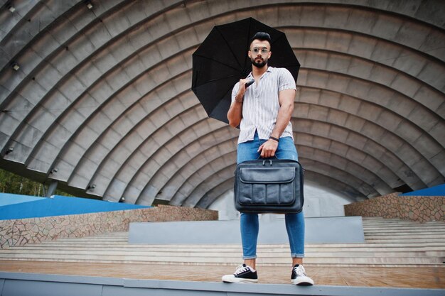 El hombre de barba árabe alto de moda usa jeans de camisa y gafas de sol con paraguas y bolsa de estuche posada en el salón de la arena